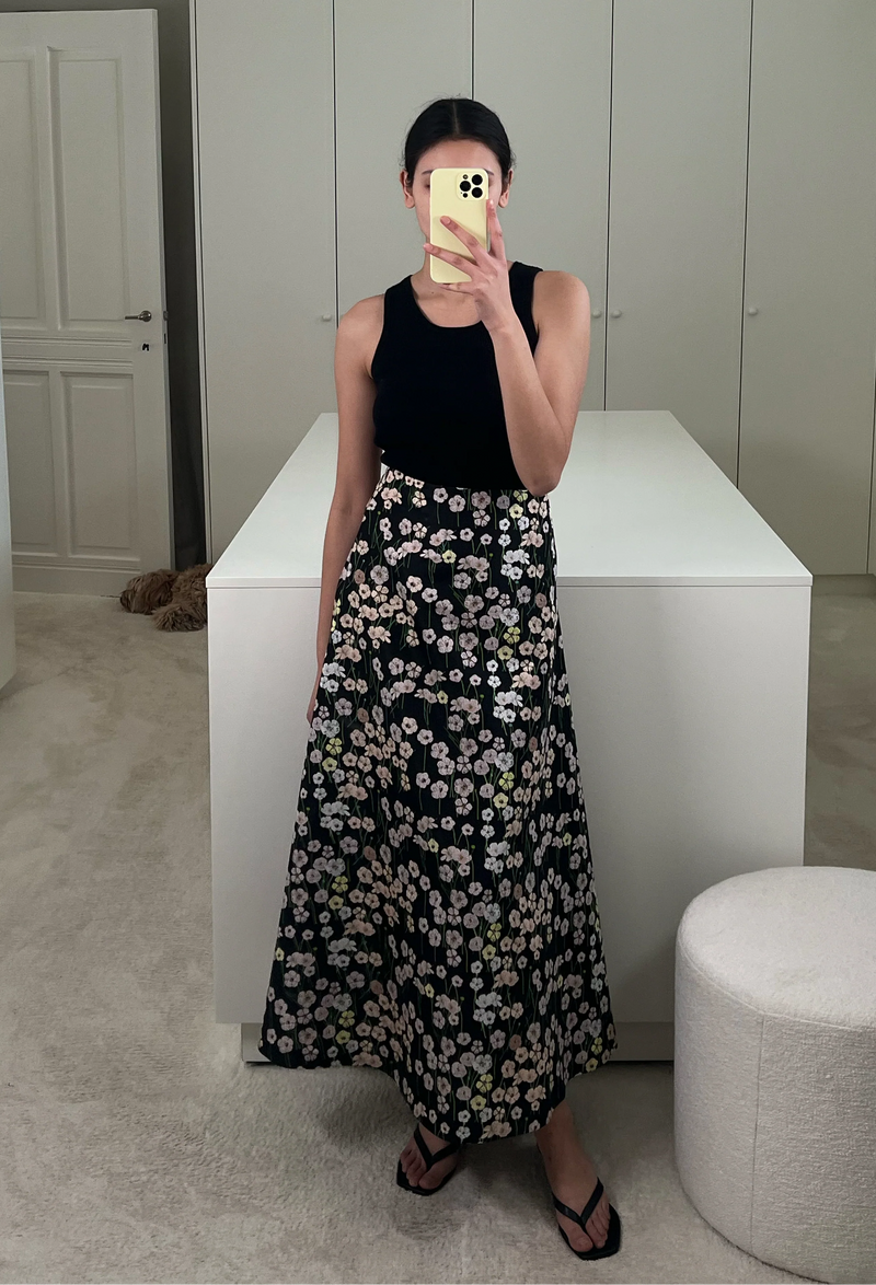 A woman in a closet taking a mirror selfie wearing a black tank top and a long black skirt with a pastel colored flowers print.