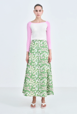 A woman wearing a pink and white long sleeve shirt and a long green skirt with a pastel colored flowers print. White background.