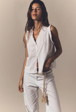 A woman wearing a sleeveless button down white top and white pants with a cord belt. Beige background.