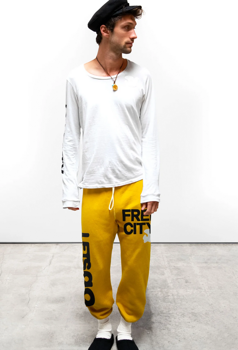 A man wearing a hat and white long sleeve tee and yellow sweatpants with free city and lets go printed in black letters. Taken on a white background.