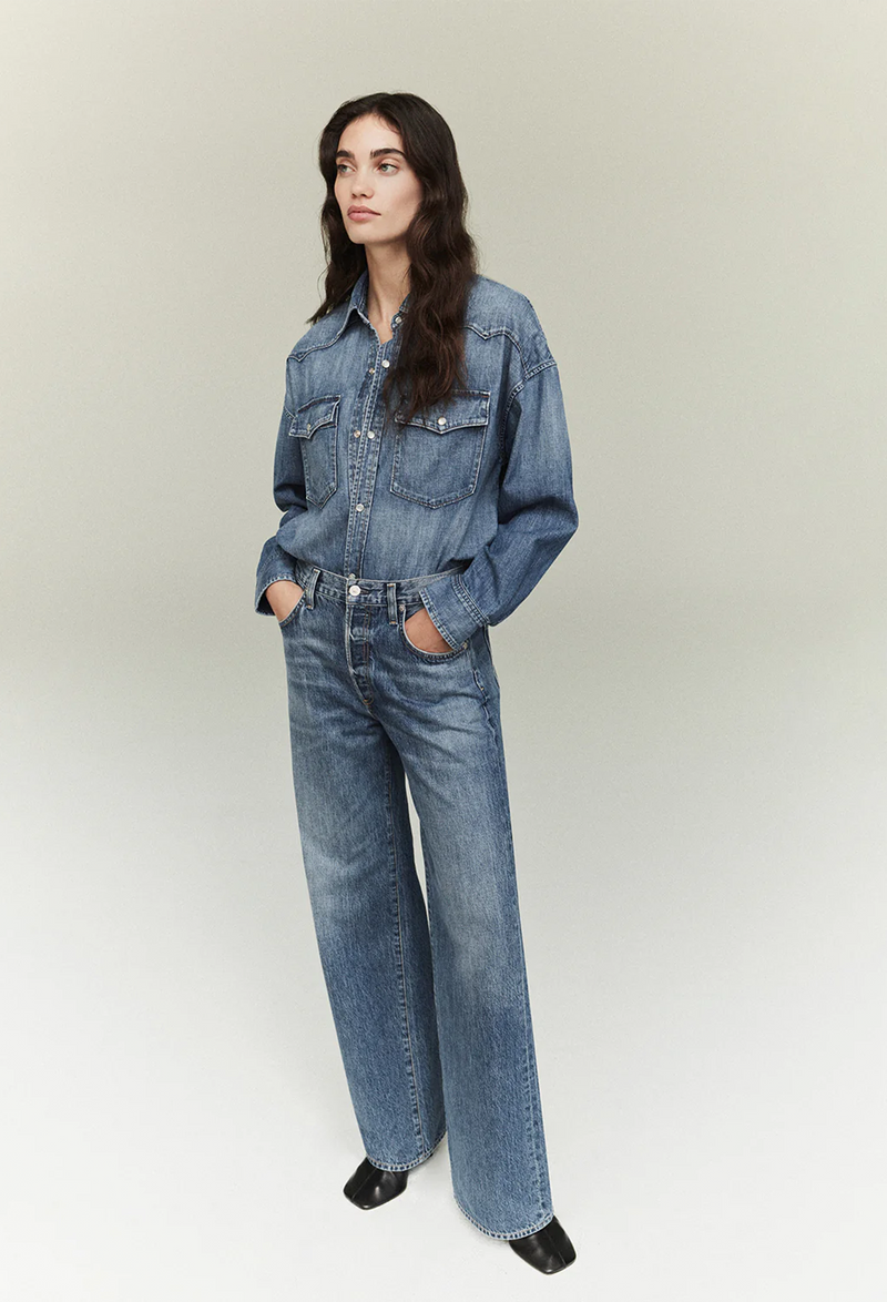 Model wears wide leg jeans with matching denim button down shirt with black shoes. 