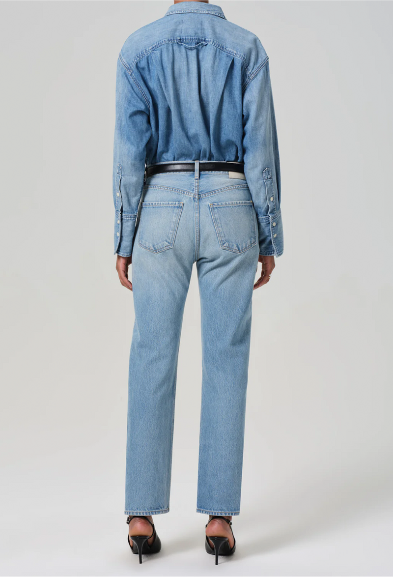From behind, model wears high rise jeans with denim button down shirt. She also wears black heels and a black belt. White background.