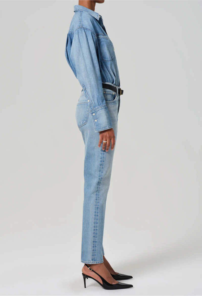 Facing the right side, model wears high rise jeans with denim button down shirt. She also wears black heels, a black belt, and a gold necklace. White background.