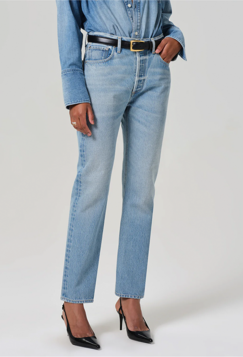 Model wears high rise jeans with denim button down shirt. She also wears black heels and a black belt. White background.