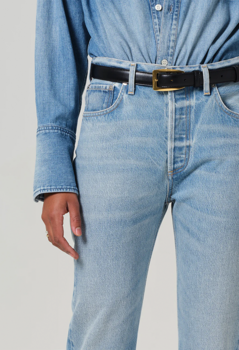 Model wears high rise jeans with denim button down shirt. She also wears black heels and a black belt. White background.
