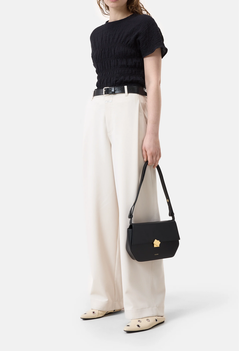 Model wears textured crinkled short sleeve black top with wide leg cream pants a black belt, white flats, and carries a black purse.
