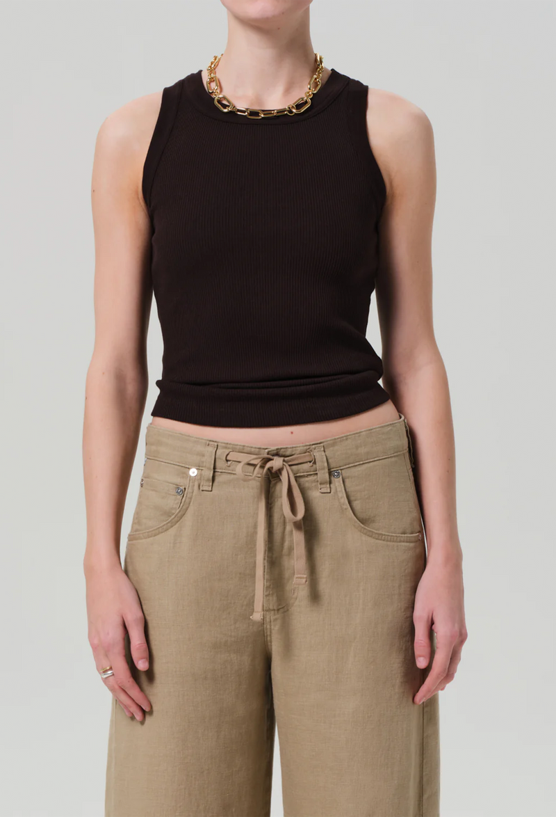 Model wears form fitting ribbed racerback chocolate brown tank. She also wears khaki drawstring pants, and a gold necklace. White background.