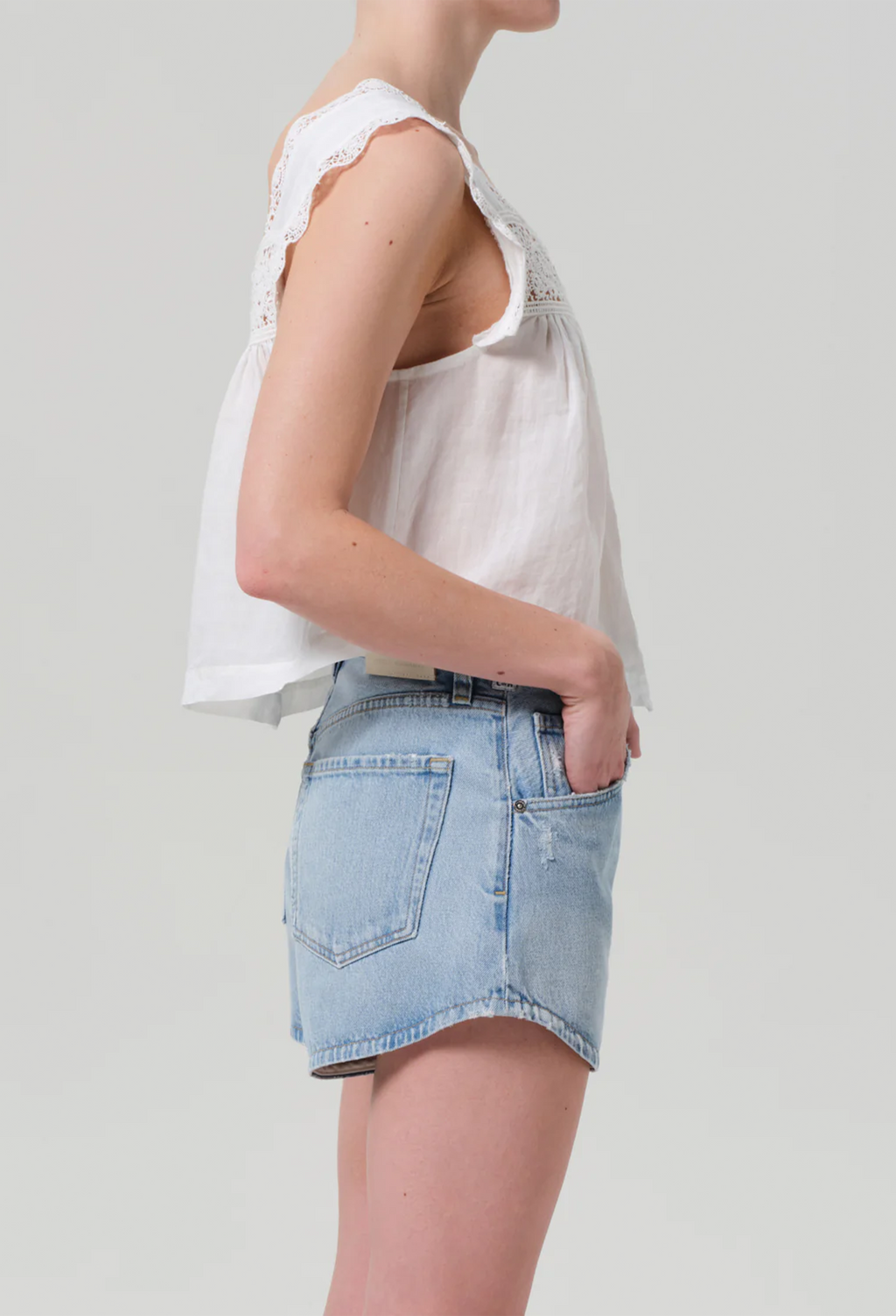 Facing the right side, model wears white square neck top with lace detailing around the front and sleeves. Top buttons up at the front. She also wears light blue jean shorts. White background.