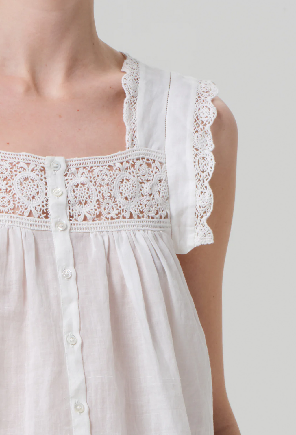 Model wears white square neck top with lace detailing around the front and sleeves. Top buttons up at the front. Photo captures her upper right side. White background.