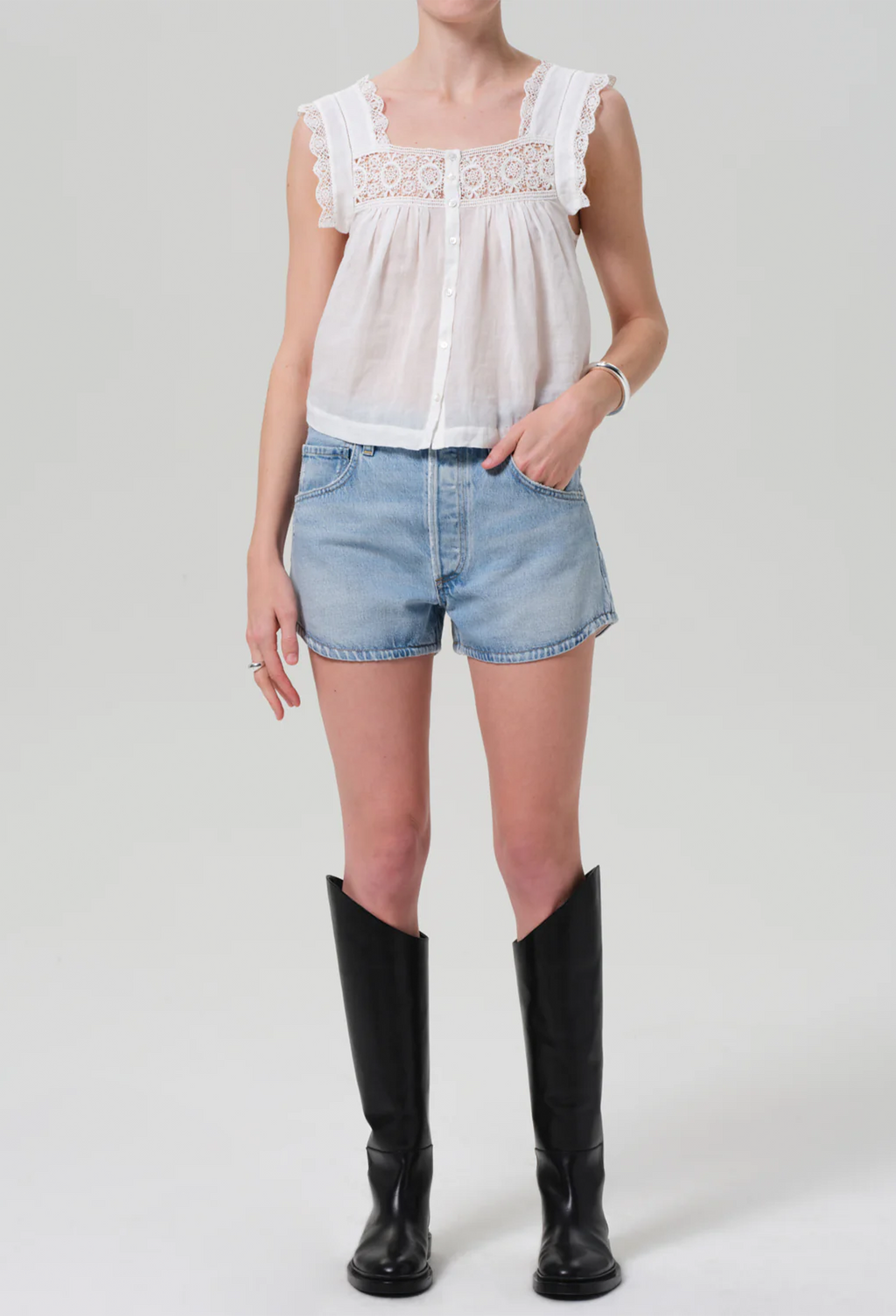 Model wears white square neck top with lace detailing around the front and sleeves. Top buttons up at the front. She also wears light blue jean shorts, and tall black boots. White background.