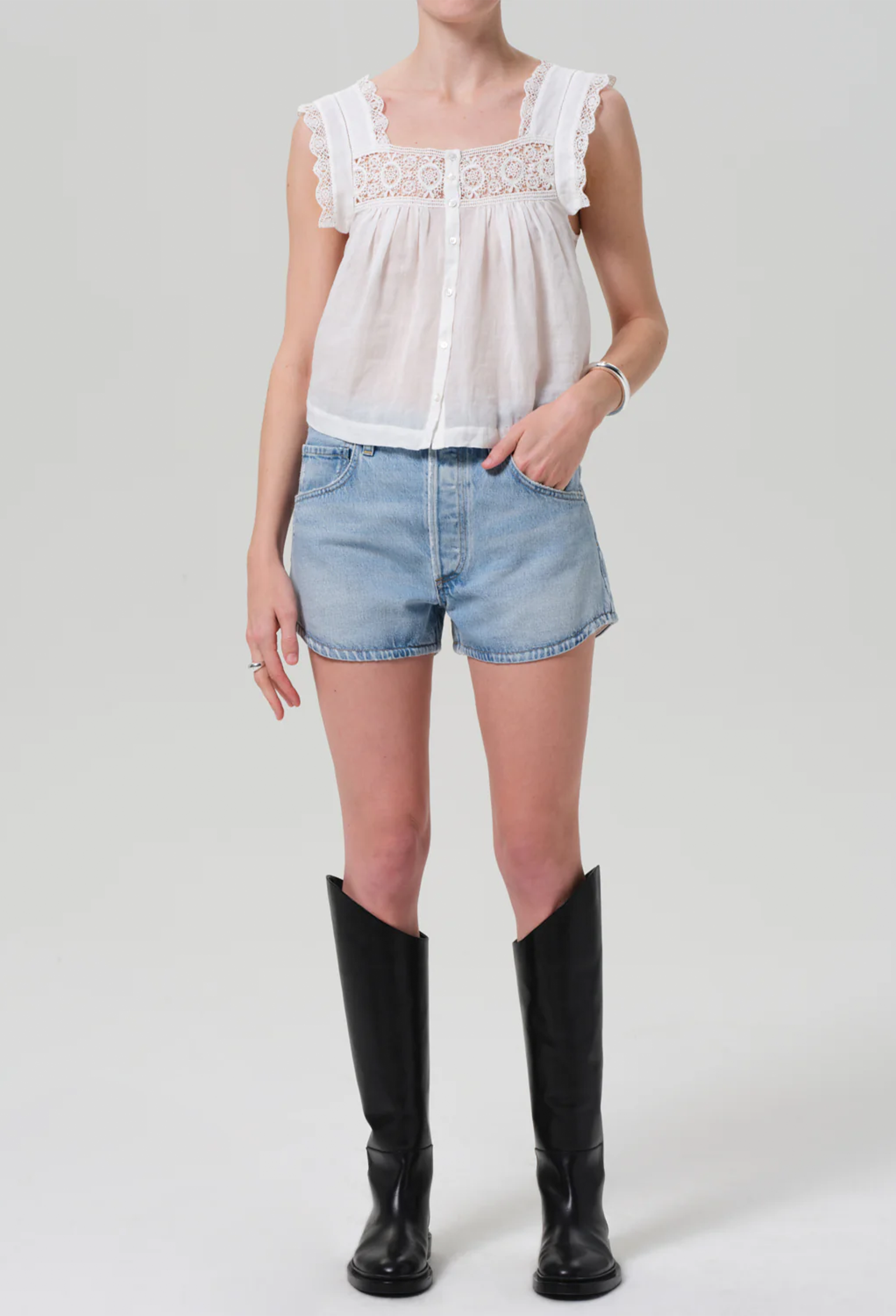 Model wears white square neck top with lace detailing around the front and sleeves. Top buttons up at the front. She also wears light blue jean shorts, and tall black boots. White background.