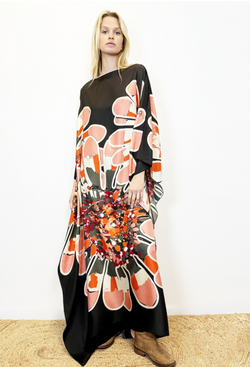 Blonde model wearing a long black silk caftan with orange and white print in the middle. Model is wearing tan distressed boots. 