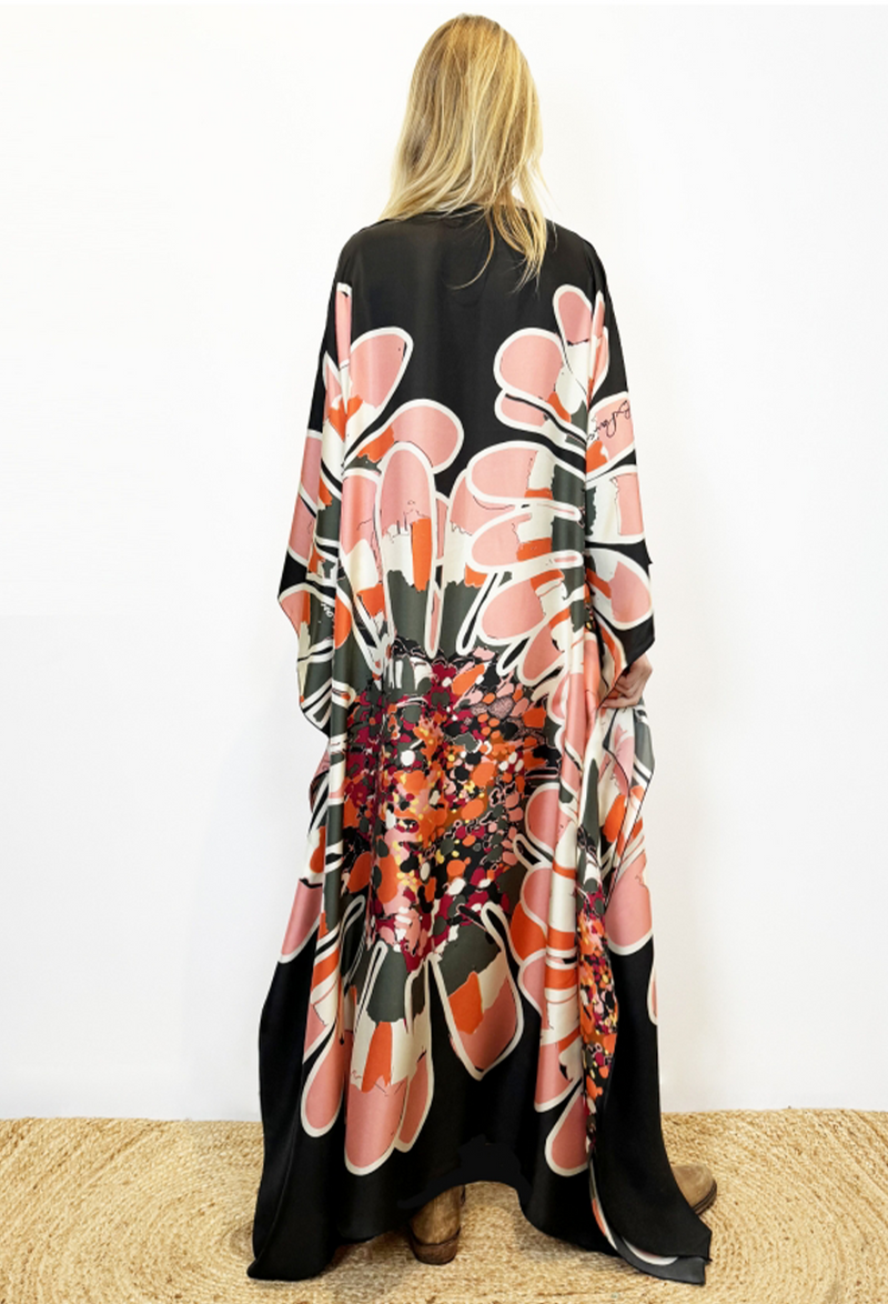 Blonde model facing white wall. She is wearing long black silk caftan that has orange and pink printing throughout. Model is also wearing distressed brown boots.