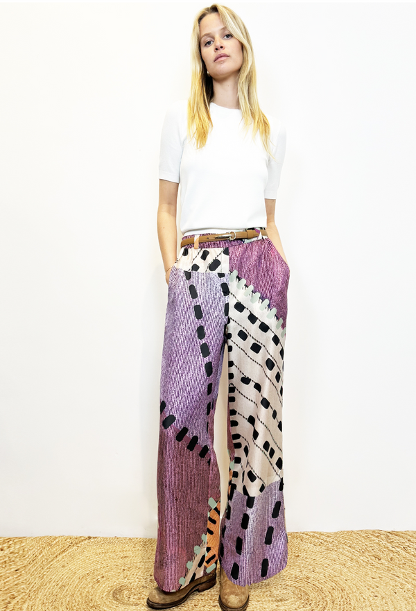 Blonde model in white T-shirt and printed purple and white silk pants with a slim brown leather belt. She is wearing brown distressed leather boots. Model is standing on carpet with white background. 