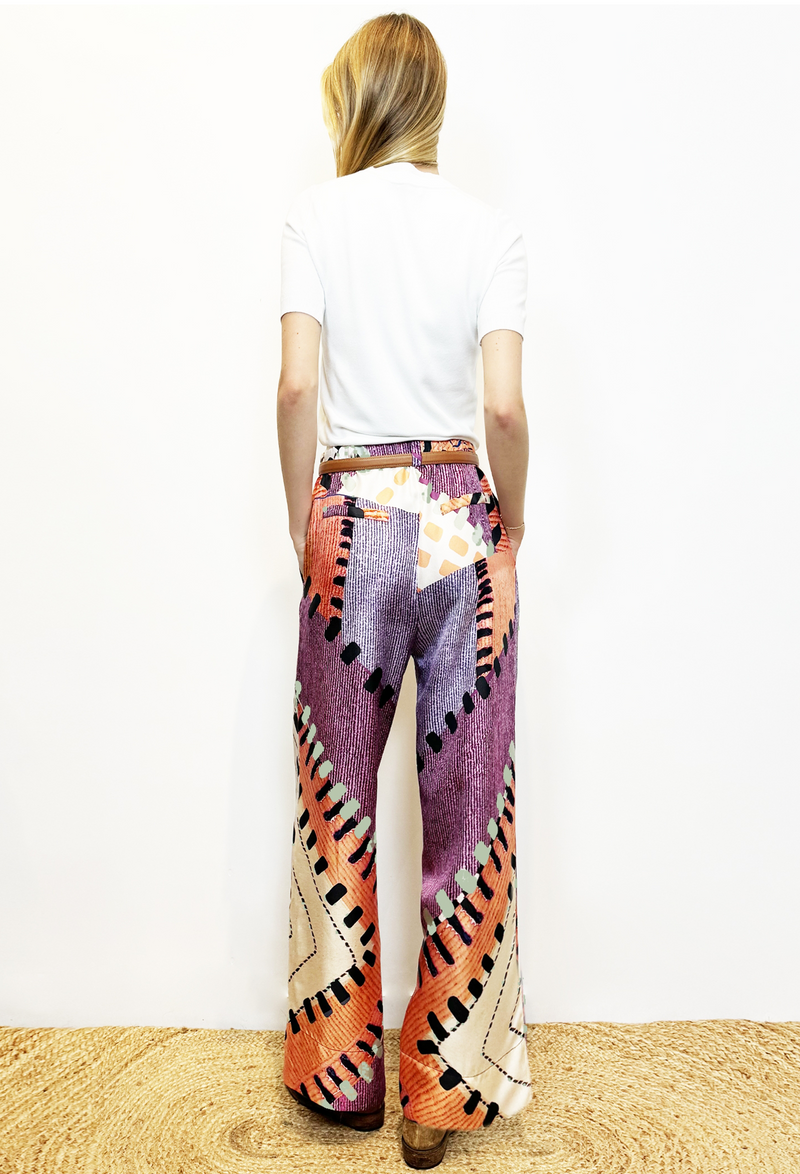 Model is facing the white wall, showing the back of the pant. Blonde model in white T-shirt and printed purple and white silk pants with a slim brown leather belt. She is wearing brown distressed leather boots. Model is standing on carpet with white background. 