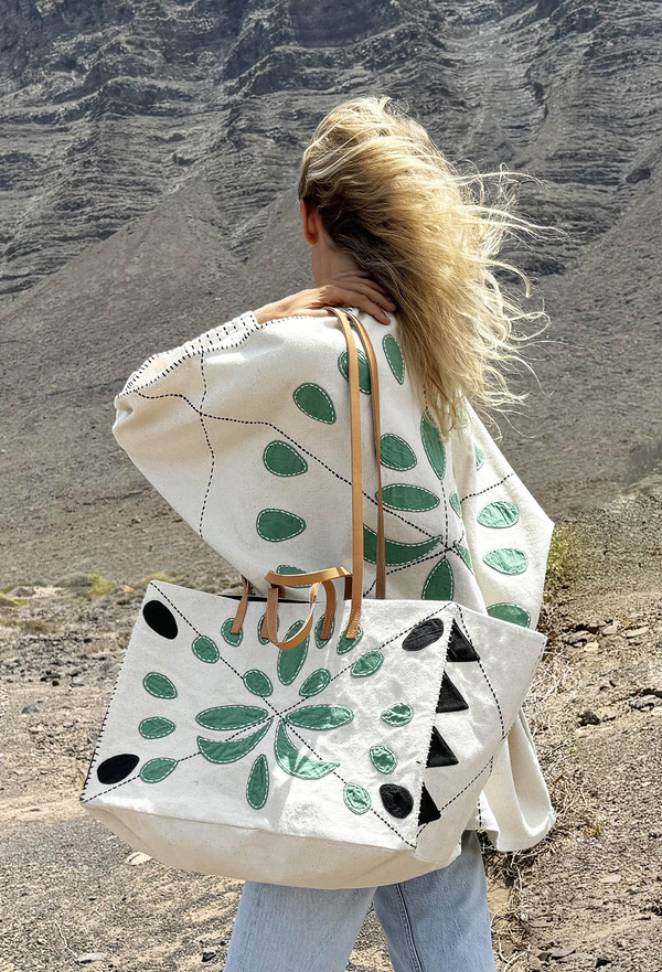 Blonde model is carrying white embroidered tote on shoulder outdoors. Tote is embroidered with green and black shapes and has both long and short tan handles.