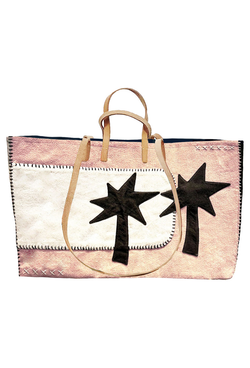 Large pink tote with stitched white patch on the left with two black palm trees overlapping. A pair of long tan handles for shoulder, and short tan handles for carrying. 