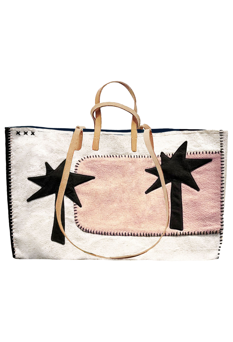 Large white tote with stitched pink patch on the left with two black palm trees on each side of the bag. A pair of long tan handles for shoulder, and short tan handles for carrying. 