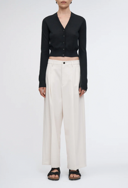 a woman wearing a cropped slim black v-neck cardigan with white pants. white background