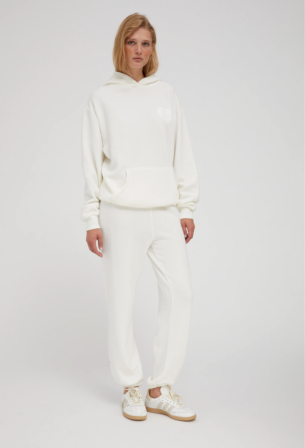 a woman wearing an off white hoodie with a white heart printed on the breast and matching sweats. white background