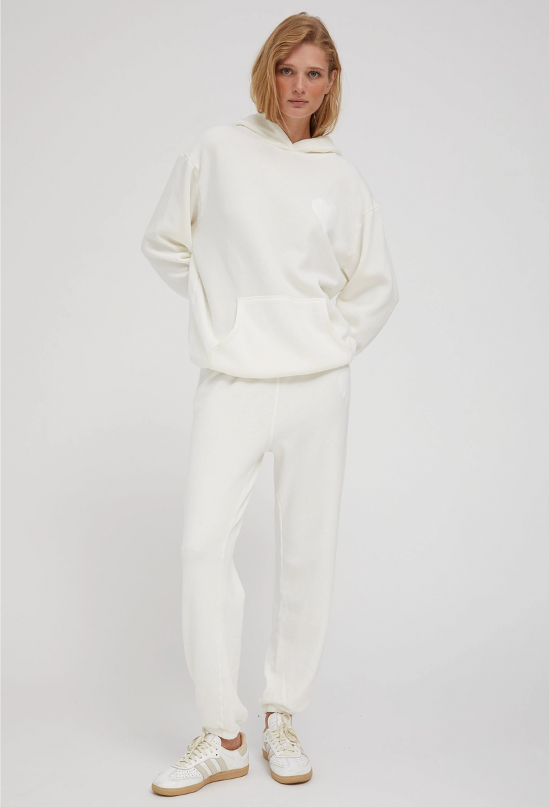 a woman wearing an off white hoodie with a white heart printed on the breast and matching sweats. white background