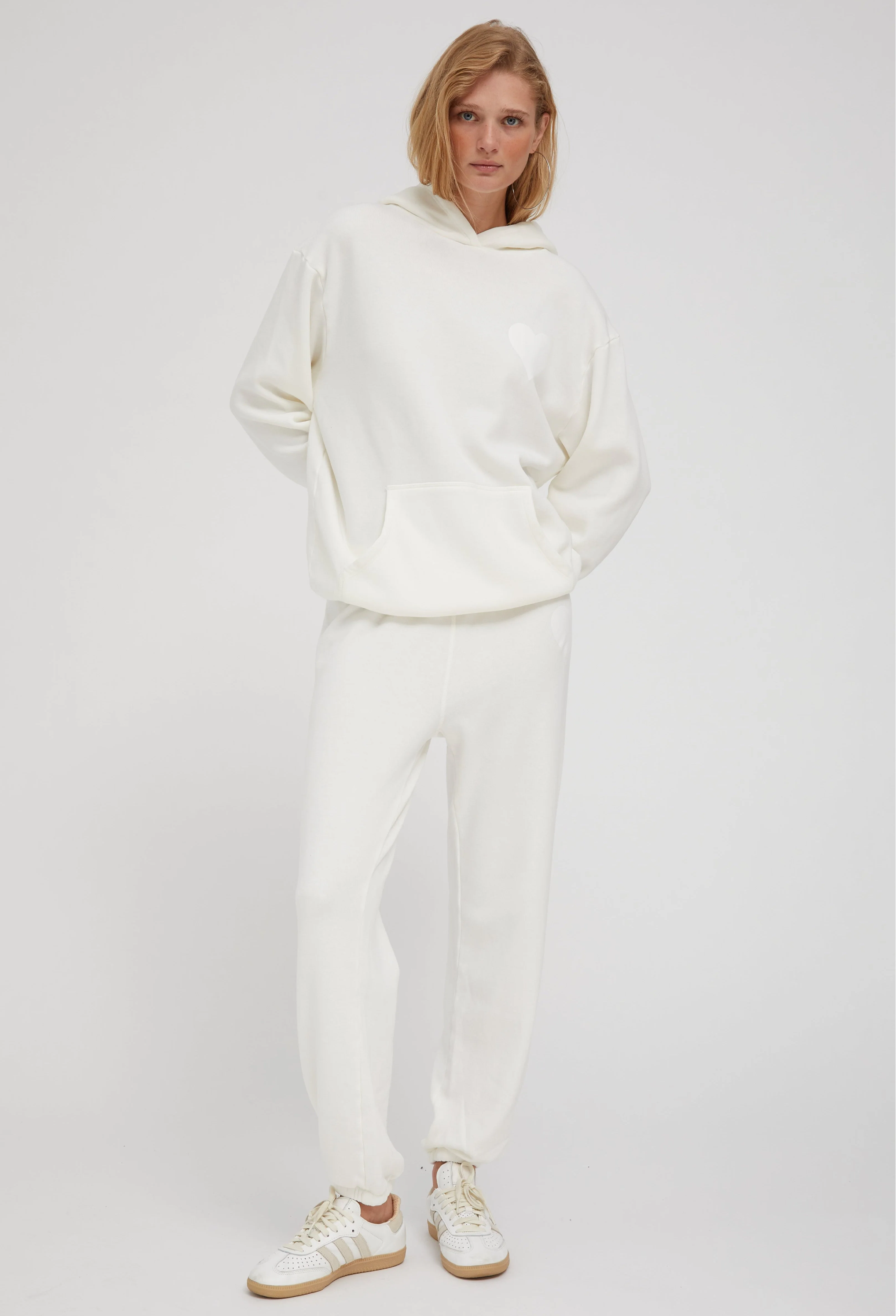 a woman wearing an off white hoodie with a white heart printed on the breast and matching sweats. white background