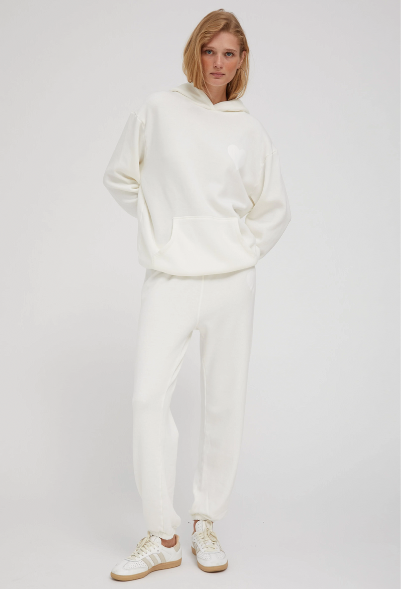a woman wearing an off white hoodie with a white heart printed on the breast and matching sweats. white background