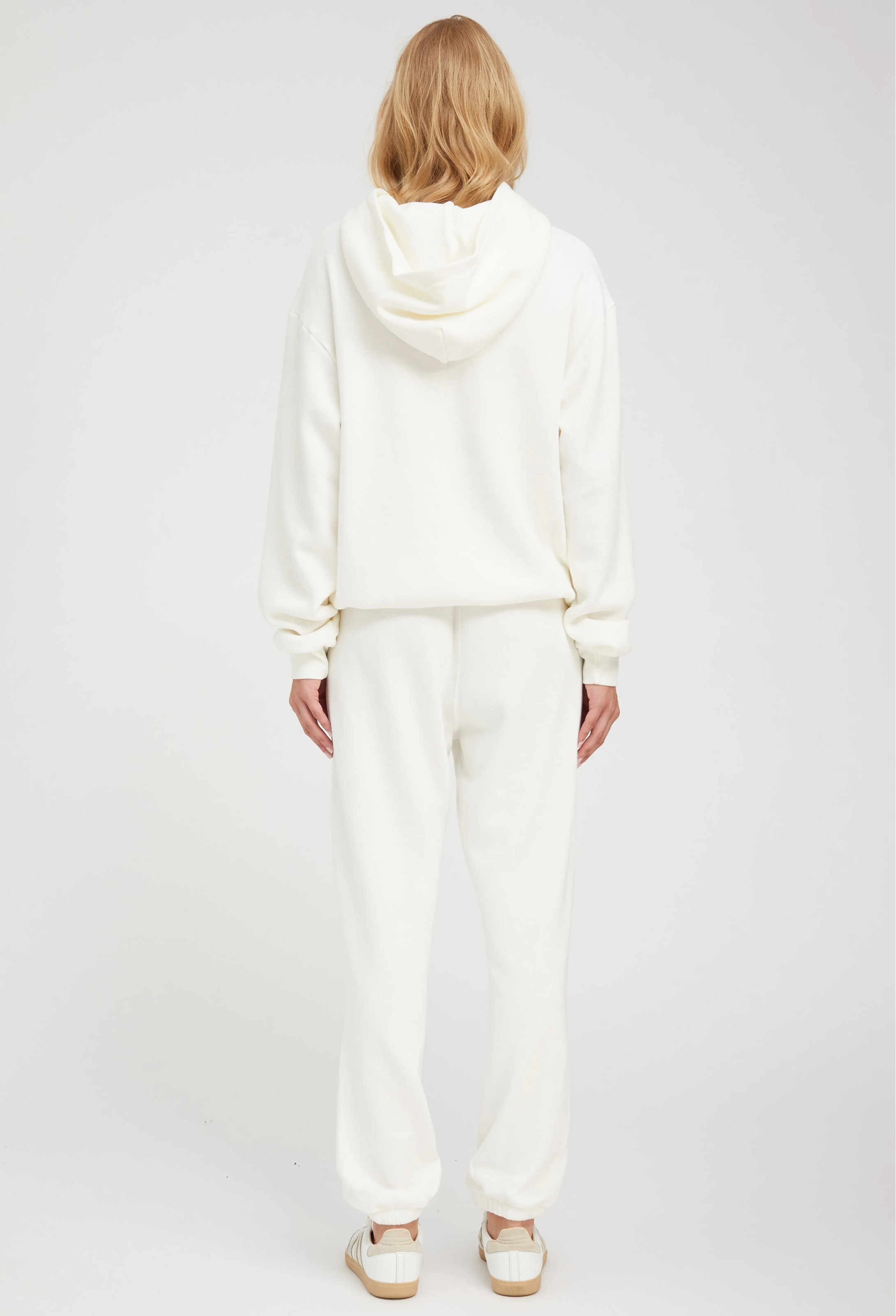 the back of a woman wearing an off white hoodie with matching sweats. white background