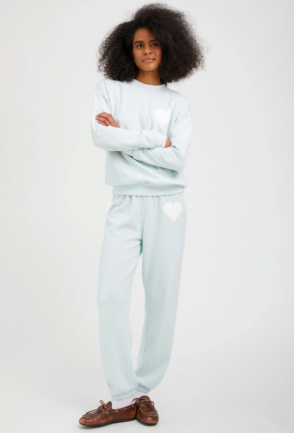 a woman wearing a pastel icy blue sweatshirt with a white heart printed on the breast with matching sweatpants. white background