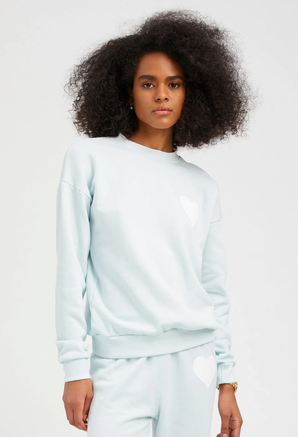 a woman wearing a pastel icy blue sweatshirt with a white heart printed on the breast with matching sweatpants. white background
