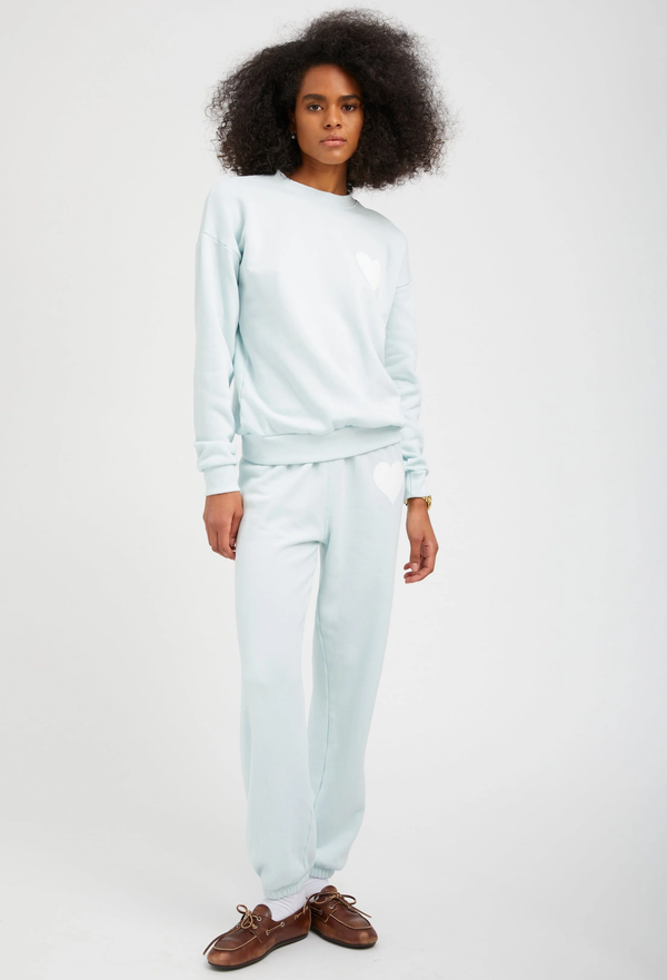 a woman wearing a pastel icy blue sweatshirt with a white heart printed on the breast with matching sweatpants. white background