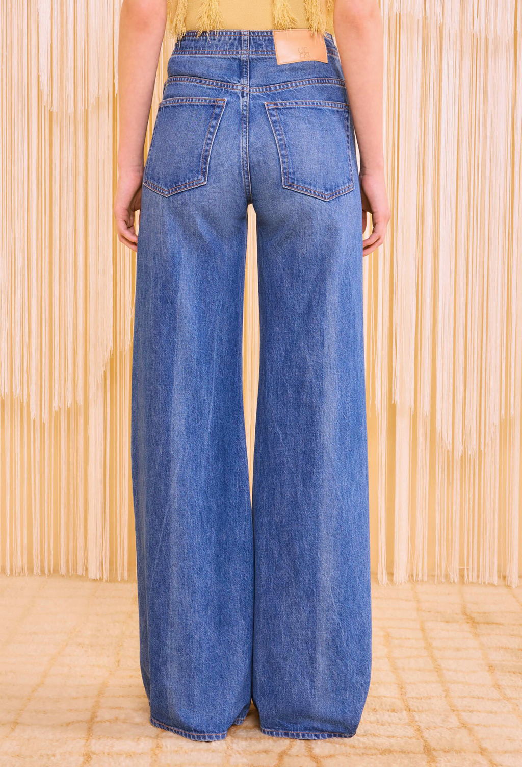 the back of a woman walking wearing a frilly yellow top and jeans with a seam down the leg. textured beige background