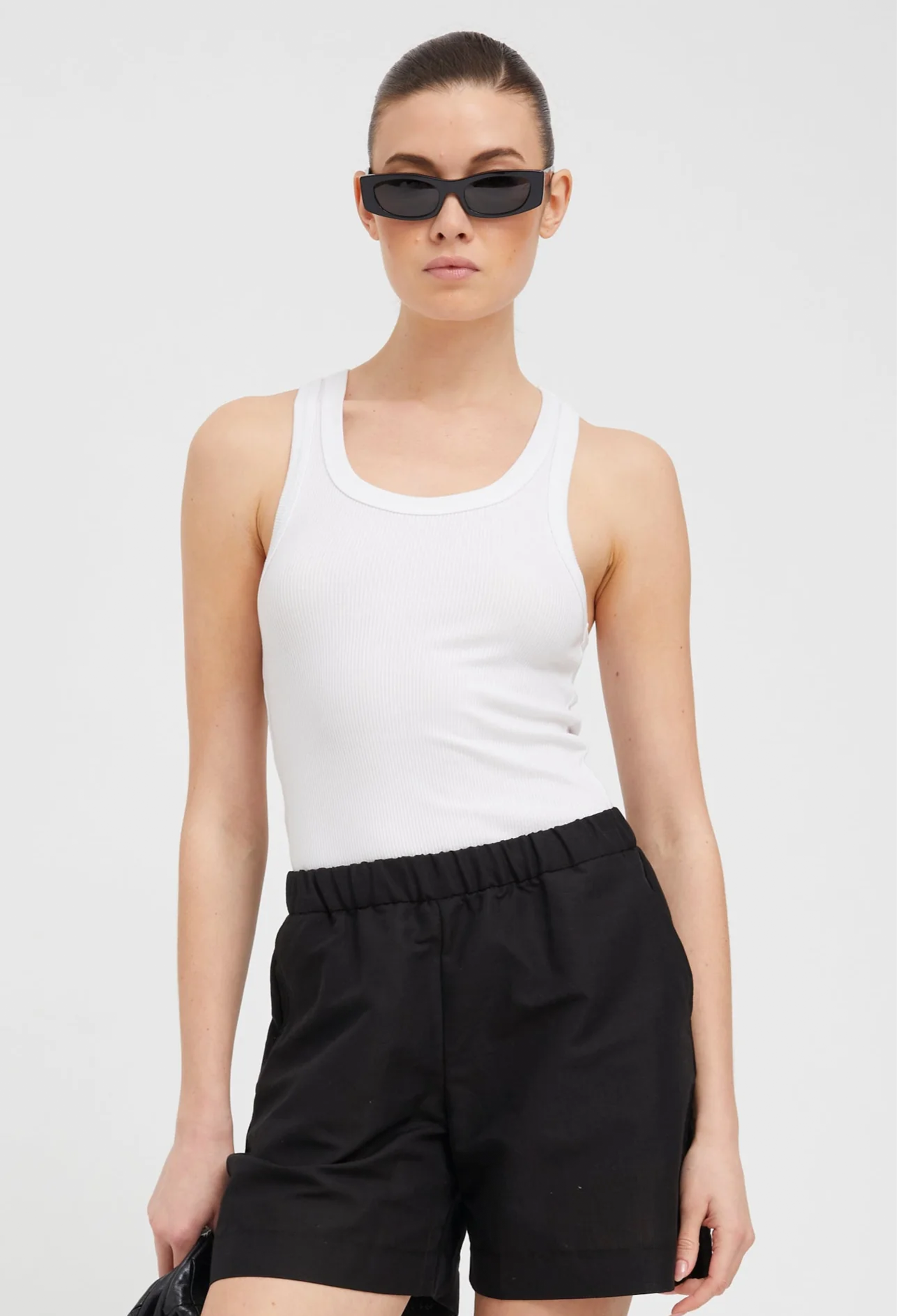 a woman wearing a scoop neck ribbed white tank top tucked into black shorts. white background
