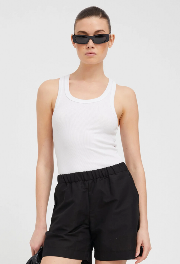 a woman wearing a scoop neck ribbed white tank top tucked into black shorts. white background