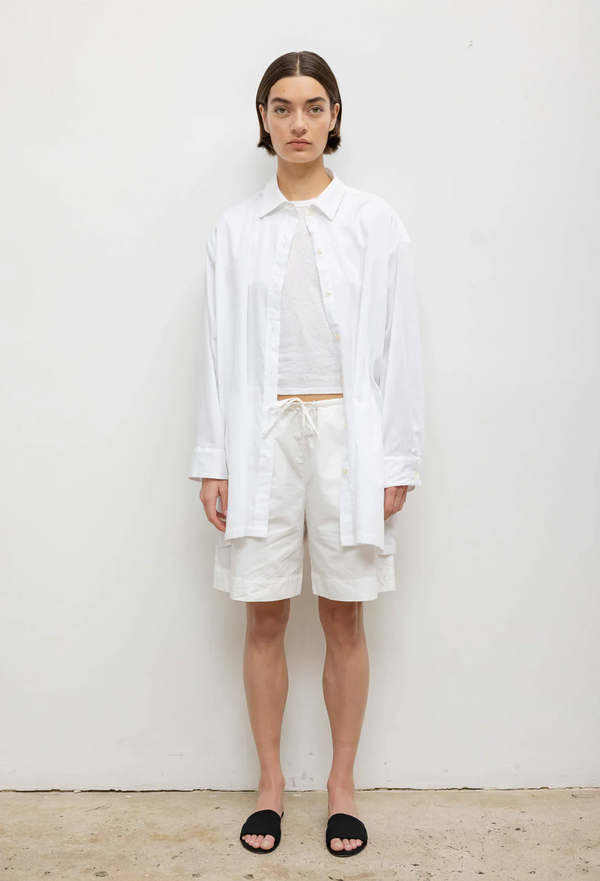 a woman wearing an oversized white button up, white t-shirt, and knee-length white drawstring shorts. white background.