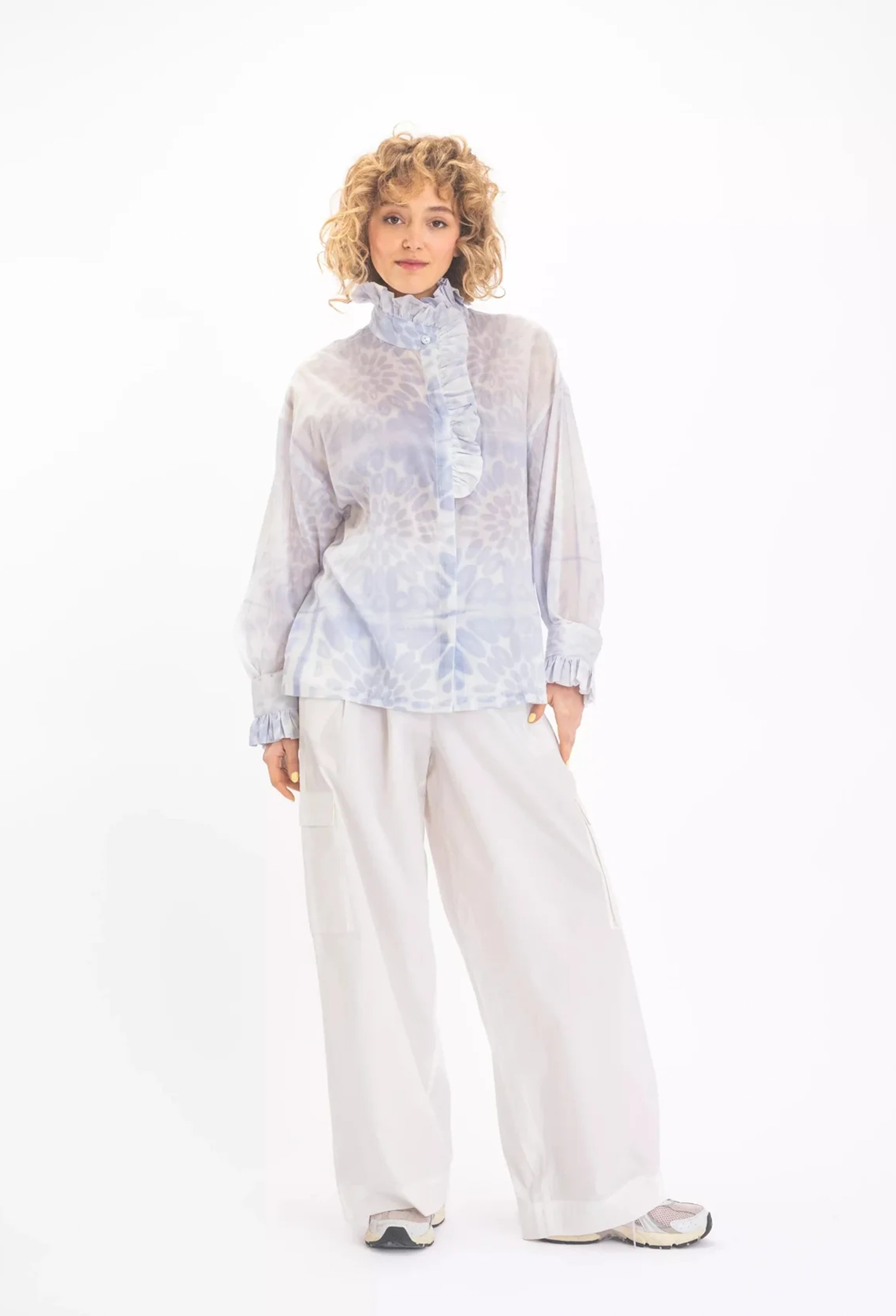 a woman wearing a white blouse with a blue floral print and a ruffled neck and sleeves. white background.