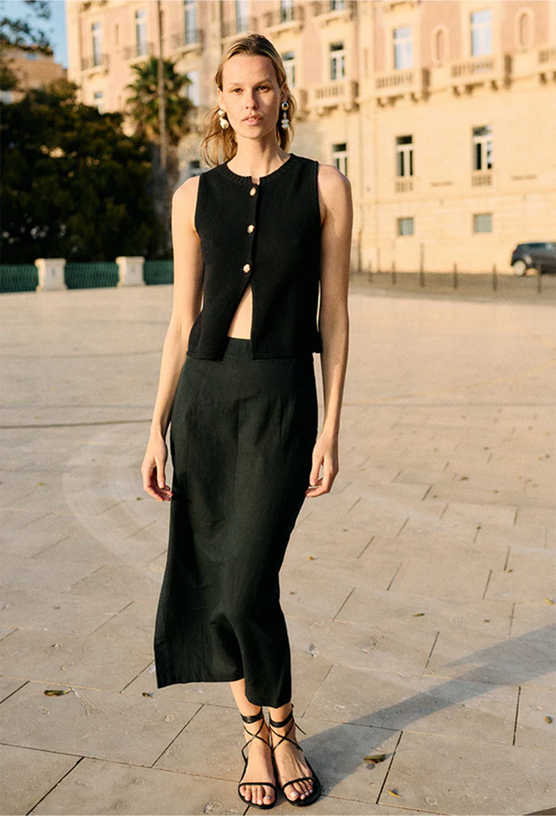 a woman outside of a large building wearing a sleeveless black knit vest with three gold buttons and a black skirt.