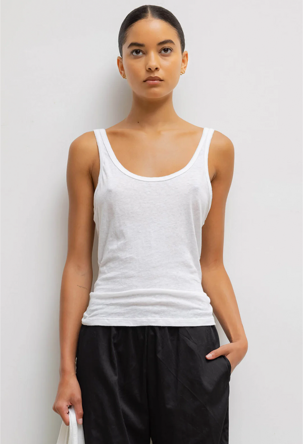 a woman wearing a white scoop neck tank top and black pants. white background.