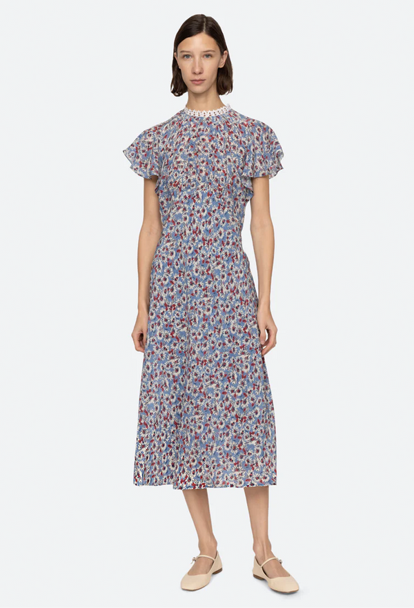 a woman wearing a blue high neck midi dress with short flutter sleeves and a white and red floral print. white background.