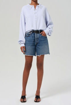 a woman wearing a light blue sweatshirt and high waisted dark wash denim shorts with a black belt. white background.