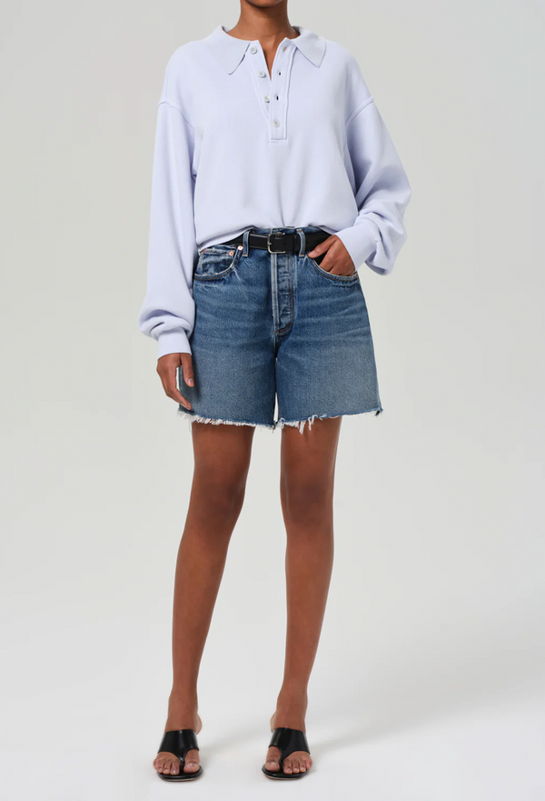 a woman wearing a light blue sweatshirt and high waisted dark wash denim shorts with a black belt. white background.