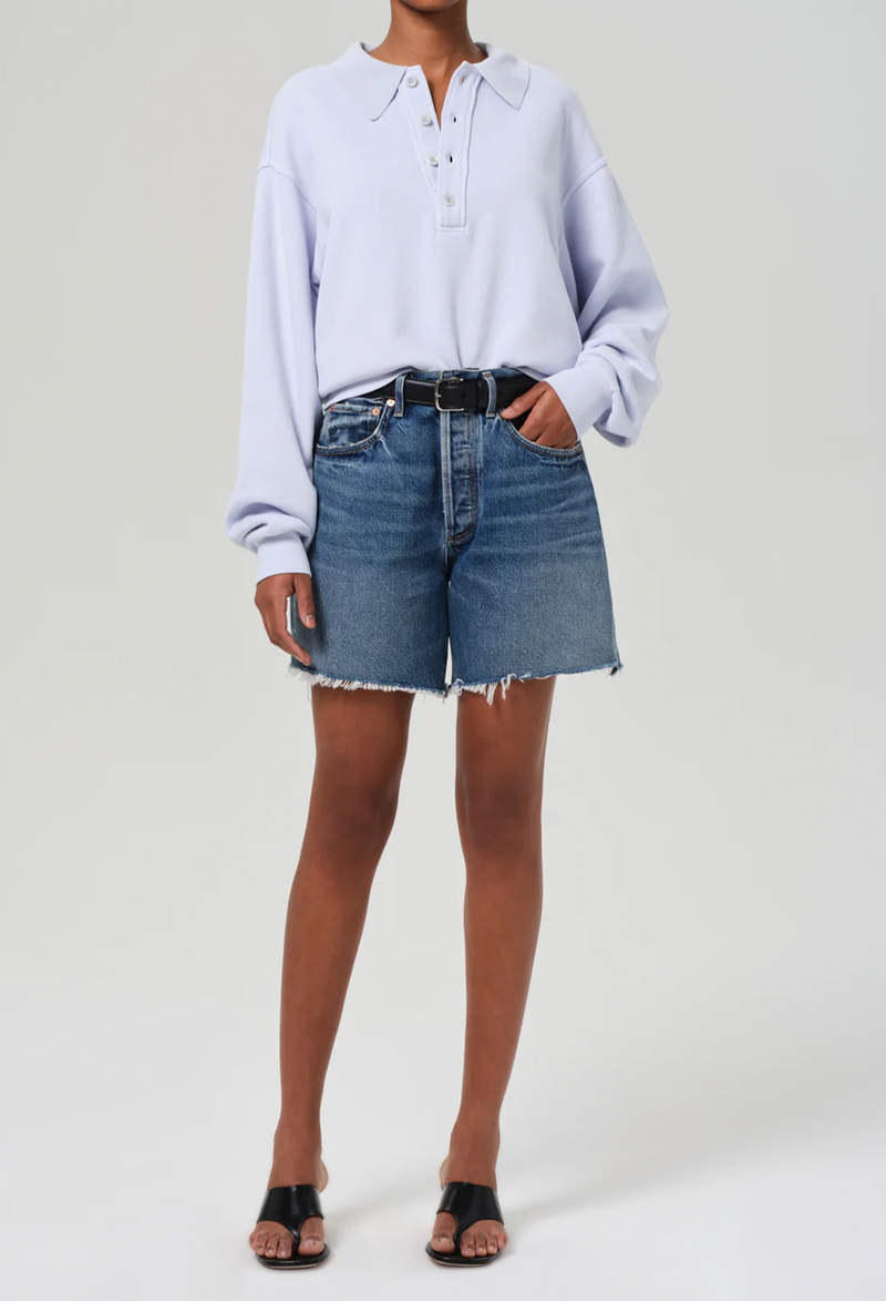 a woman wearing a light blue sweatshirt and high waisted dark wash denim shorts with a black belt. white background.