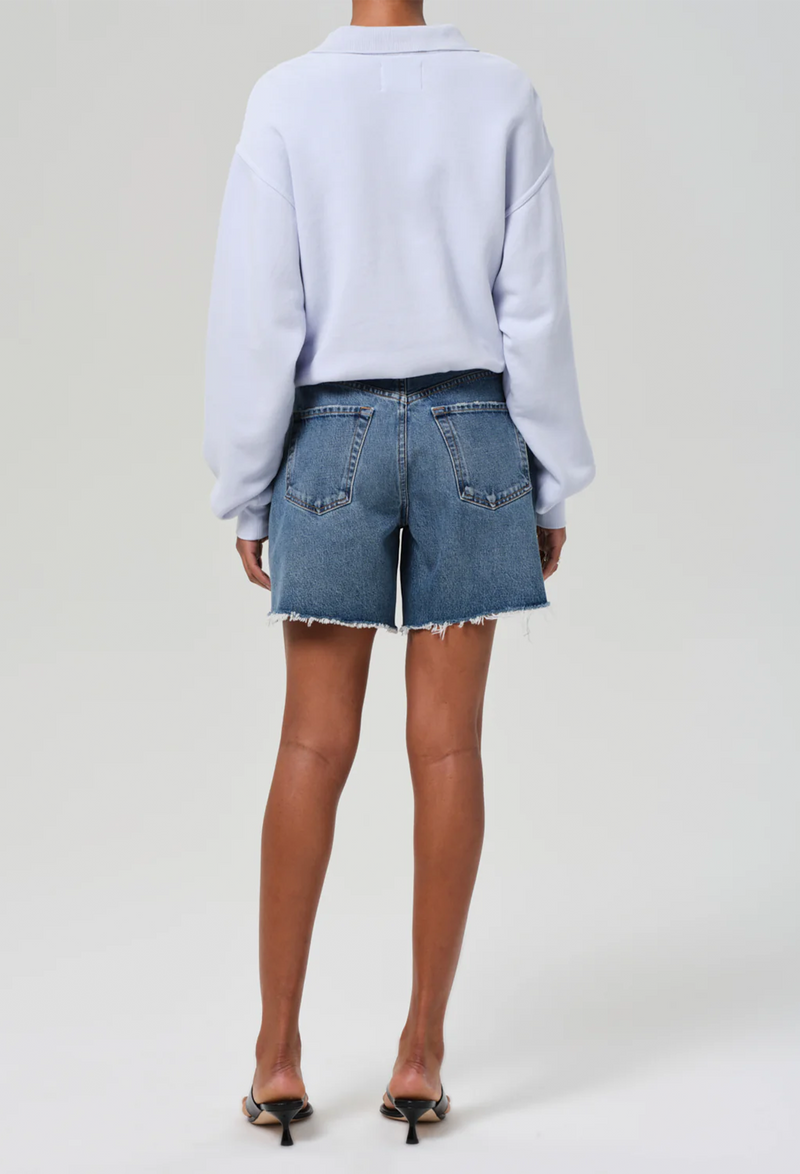 the back of a woman wearing a light blue sweatshirt and high waisted dark wash denim shorts. white background.