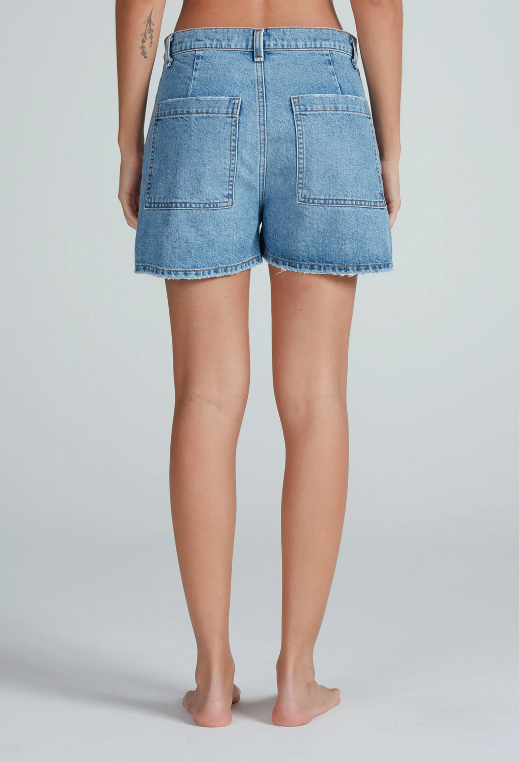 the back of a woman wearing light denim shorts with two large patch pockets. white background.