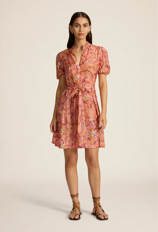 a model wearing a coral mini dress with a tie waist, flared skirt, short puff sleeves, high neck with loop closure, and a yellow, red, and orange floral pattern. white background.