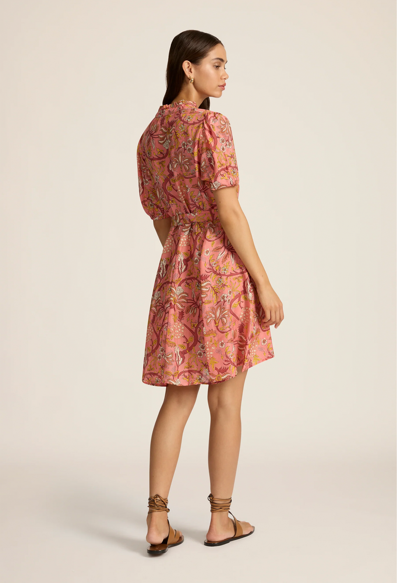 the back of a model wearing a coral mini dress with a tie waist, flared skirt, short puff sleeves, high neck with loop closure, and a yellow, red, and orange floral pattern. white background.