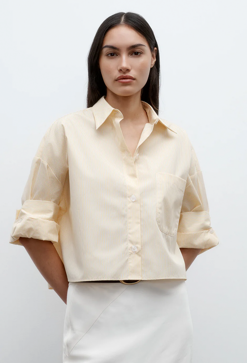 a model wearing a white and yellow striped cropped button up shirt with rolled sleeves and white bottoms. white background.