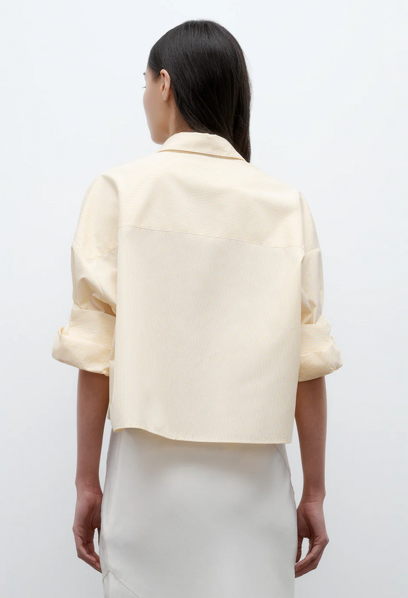 the back of a model wearing a white and yellow striped cropped button up shirt with rolled sleeves and white bottoms. white background.