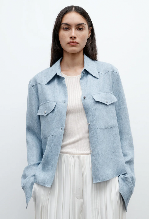 a model wearing a light blue cropped button up work shirt with two chest flap pockets with a white shirt and white pants. white background.