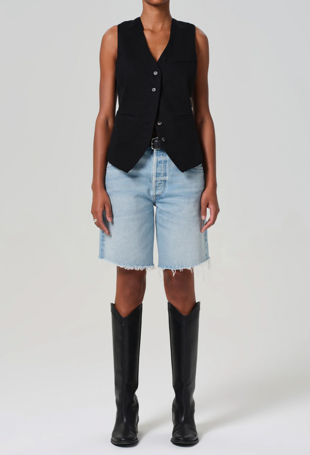 a model wearing a black vest and long light wash denim shorts with a frayed hem. white background.